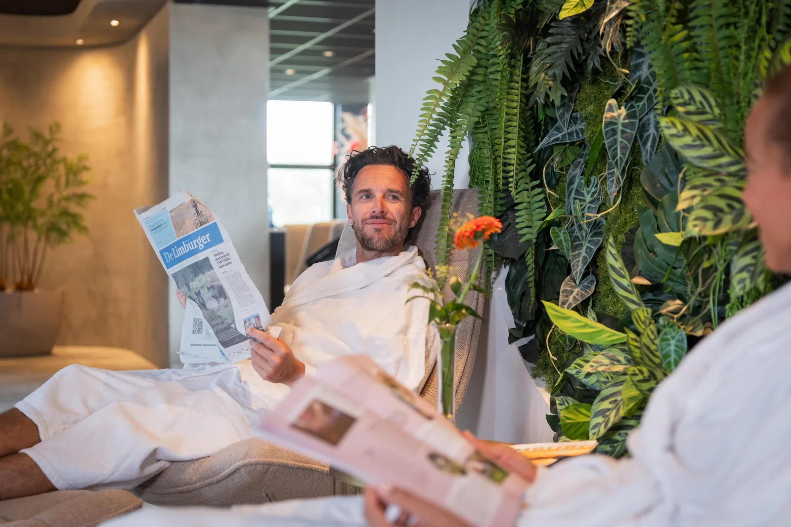 Ein Mann und eine Frau in weißen Bademänteln sitzen auf hellen Liegen im Ruhebereich, er liest eine Zeitung, sie blättert in einer Zeitschrift, dahinter eine Wand aus grünen Pflanzen